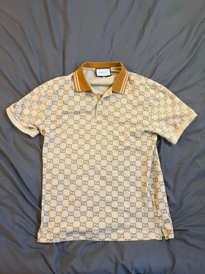 Beige Gcci pikétröja med monogram - Snygg beige pikétröja från Gcci med klassiskt G-monogram över hela plagget. Kragen är i kontrasterande brunt med vita ränder. Tröjan har korta ärmar och knäppning vid halsen. Perfekt för dig som gillar exklusiv streetstyle.