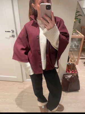 Vinröd cape-kappa med knappar - Säljer en vinröd cape-kappa med knappar framtill och hög krage. Kappan har en lös och vid passform med vida ärmöppningar, perfekt för lager-på-lager. Materialet ser ut att vara ullblandning och den har en stilren, minimalistisk look. St 38