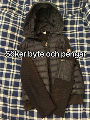 Svart cardigan  med huva från Moncler - Snygg svart dunjacka från Moncler med stickade ärmar och muddar. Jackan har en stor huva, dragkedja framtill och en ficka med dragkedja på ena ärmen. Perfekt för kalla dagar och riktigt stilren design.