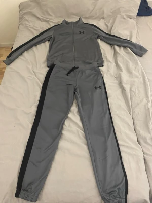 Grå träningsset från Under Armour - Säljer ett grått träningsset från Under Armour med svarta detaljer längs ärmar och ben. Setet består av en zip-jacka och matchande byxor med resår i midja och benslut. Perfekt för träning eller chill. Materialet är mjukt och följsamt.