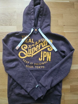 Lila-brun hoodie från Superdry ( Stl. S men sitter som mer som en M)  - Säljer en lila hoodie från Superdry med stor gul logga och japansk text på bröstet. Hoodien har vit dragsko, känguruficka och ribbade muddar. Perfekt för dig som gillar streetwear och vill ha något bekvämt och snyggt. Är en S men sitter med som en Storlek M 