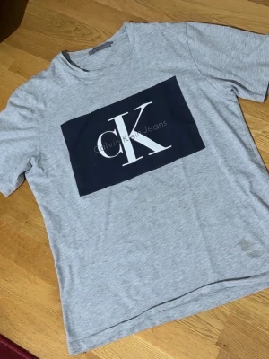 Grå Calvin Klein t-shirt - Snygg grå t-shirt från Calvin Klein Jeans med stort svart tryck och vit CK-logga på bröstet. Klassisk passform och korta ärmar. Perfekt för dig som gillar stilrena och trendiga plagg med tydlig logga. Tillverkad i mjuk bomull för skön känsla. Litet fläck på sidan av tröjan annars fint skick.