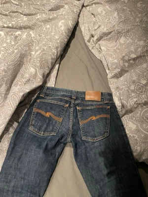 Blå jeans från Nudie Jeans - Säljer ett par klassiska blå jeans från Nudie Jeans med raka ben och snygga kontrastsömmar på bakfickorna. Jeansen har en läderpatch bak i midjan och är tillverkade i slitstark denim. Perfekta för en avslappnad och trendig look. 