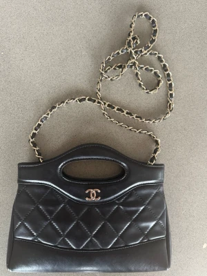 Quilitat väska i läder - Elegant svart handväska med quiltat mönster på framsidan och stilren design. Väskan har ett integrerat handtag upptill samt en avtagbar eller nedfällbar kedjerem i metall för flexibel användning – kan bäras både i handen och över axeln. Framsidan pryds av en diskret metalllogotyp som ger ett sofistikerat uttryck.  Insidan är helfodrad i mörkt tyg och försedd med en innerficka för mindre tillhörigheter. Väskan stängs med magnetknapp och har en kompakt men praktisk storlek, lämplig för vardag elle