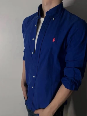 Blå skjorta från Ralph Lauren - Tjo Boss! Säljer en snygg blå skjorta från Ralph Lauren med klassisk krage och knappar framtill. Custom fit-modell i bomull med lång ärm och ikonisk röd broderad logga på bröstet. Den är knappast använd och i ett extremt gott skick, storlek 18 motsvarar då storlek S kan gå ner i pris