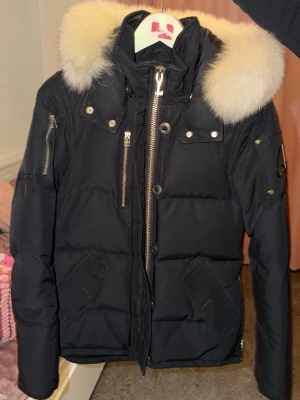 Svart dunjacka med pälskrage Moose Knuckles - Snygg svart dunjacka från Moose Knuckles med stor vit pälskrage och flera dragkedjor. Jackan har huva, två fickor fram och detaljer i silver. Perfekt för kalla vinterdagar och riktigt trendig stil.