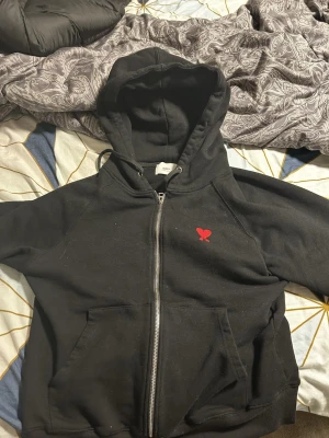 Svart hoodie med röd Ami de Coeur - Säljer en svart hoodie med dragkedja från AMI Paris. På bröstet finns det ikoniska röda Ami de Coeur-broderiet. Hoodien har huva med dragsko och två fickor framtill. Tillverkad i mjuk bomull för maximal komfort. Nypris ca 4 tusen men säljer för endast 2,5 tusen