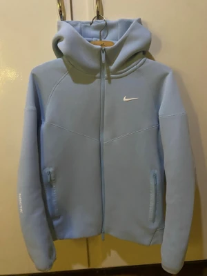 Ljusblå Nike NOCTA hoodie - Snygg ljusblå hoodie från Nike NOCTA med dragkedja framtill och två fickor med dragkedja. Har en vit Nike-logga på bröstet och NOCTA-tryck på ärmen. Mjuk och skön med huva och coola sömdetaljer. Perfekt för en avslappnad streetwear-look.
