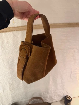 Brun mocka handväska med spänne arket  - Säljer en snygg och populär handväska i brun mocka med bred axelrem och dekorativt spänne på sidan. Väskan har en mjuk, fyrkantig form och är perfekt för dig som gillar stilrena detaljer och jordnära färger.