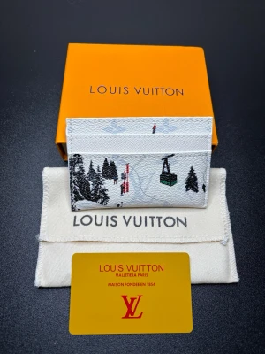 Louis Vuitton skidmotiv kortfodral - Snyggt kortfodral från Louis Vuitton i vitt läder med vinterlandskap, skidåkare och liftar som motiv. Diskret LV-monogram i bakgrunden. Kompakt och stilren design med flera kortfack. Perfekt accessoar för dig som gillar exklusiva detaljer och unik design.