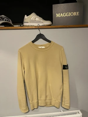 Beige sweatshirt från Stone Island - Säljer en beige sweatshirt från Stone Island med klassisk rund hals och den ikoniska svarta logotypen på vänster ärm. Tröjan har en clean look med ribbade muddar vid ärmslut och nederkant. Perfekt för dig som gillar streetwear och stilren design.
