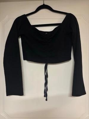 Svart croppad topp med snörning - Trendig svart croppad topp med långa ärmar och snygg snörning framtill. Toppen har en djup v-ringning och är tillverkad i ett ribbat material som ger en skön passform. Perfekt för att lyfta din outfit med en edgy touch.