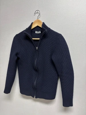 Mörkblå ziptröja från Gran Sasso - Mörkblå stickad ziptröja från Gran Sasso, tillverkad i Italien. Tröjan har hög krage med ribbad kant, hel dragkedja framtill och ribbade muddar vid ärmslut och nederkant. Perfekt för lager på lager och snygg till jeans. Enda nackdel är att dragkedjan kan strula lite ibland och den grejen man håller i när man drar är av på den övre men det går fortfarande bra.