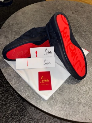 Blå sneakers från Christian Louboutin - Snygga svarta sneakers från Christian Louboutin i mocka med klassisk röd sula och diskret logga på sidan. Skorna har svarta skosnören och en låg profil som ger en clean och exklusiv look. Perfekta för dig som vill sticka ut med stil.