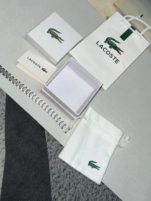 Silverfärgat armband från Lacoste - Snyggt armband i silverfärgad metall från Lacoste med länkdesign och ingraverade detaljer på varje länk. Armbandet har en stilren och modern look med Lacostes ikoniska krokodillogga på spännet. Levereras med originalask, påse och presentpåse.