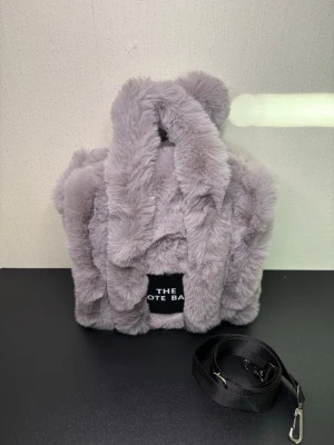 Grå fluffig handväska med axelrem - Supersöt handväska i mjuk, grå fuskpäls med korta handtag och avtagbar svart axelrem. Väskan har en fyrkantig form och en liten svart patch med texten 'THE TOTE BAG' framtill. Perfekt för dig som gillar trendiga accessoarer.