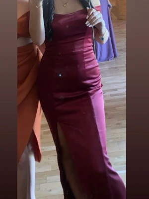 Vinröd balklänning  - Snygg vinröd långklänning i figurnära modell med axelbandslös design och hög slits framtill. Klänningen har en elegant, enkel look och är tillverkad i ett glansigt material som ger en lyxig känsla.