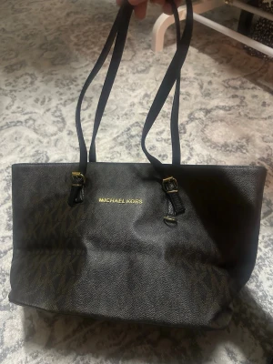 Svart handväska från Michael Kors - Helt ny Snygg svart handväska från Michael Kors med guldiga detaljer och justerbara axelband. Väskan har ett diskret MK-mönster och är tillverkad i tåligt konstläder. Perfekt för dig som vill ha en stilren och rymlig väska med exklusiv känsla.