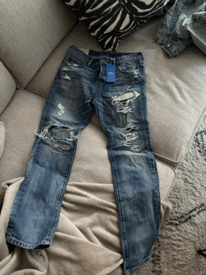 Blå slitna jeans Abercrombie & Fitch - Säljer ett par blå jeans från Abercrombie & Fitch med kraftiga slitningar och hål på både fram- och baksida. Jeansen har klassisk femficksmodell och raka ben. Tillverkade i denim med snygg tvätt och slitna detaljer för en trendig look.