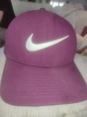 Lila keps från Nike med vit logga - Snygg lila keps från Nike med den klassiska vita swoosh-loggan broderad framtill. Kepsen har böjd skärm och justerbar passform. Tillverkad i 100% bomull för en bekväm känsla. Perfekt accessoar för en sportig och trendig look.