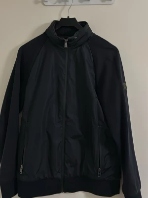 Svart vindjacka med dragkedja - Hugo boss vindjacka med hög krage och hel dragkedja framtill. Storlek S men stor i storlek så kan passa M. Jätte bra skick inte mycket använd. Nypris 1700kr