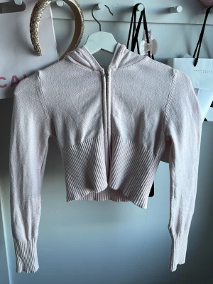 Ljusrosa cropped hoodie med dragkedja - Snygg ljusrosa cropped hoodie med dragkedja framtill och ribbade detaljer vid midjan och ärmsluten. Hoodien har långa ärmar och en klassisk huva. Perfekt för en chill och trendig look. Materialet är mjukt och stickat för extra komfort.