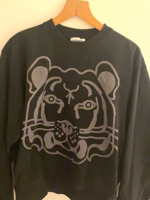 Svart sweatshirt med tigertryck Kenzo - Cool svart sweatshirt från Kenzo med stort tigertryck i grått på framsidan och en liten tigerpatch bak i nacken. Tröjan har rund halsringning och ribbade muddar vid ärmslut och nederkant. Perfekt för dig som gillar streetwear och statementplagg.