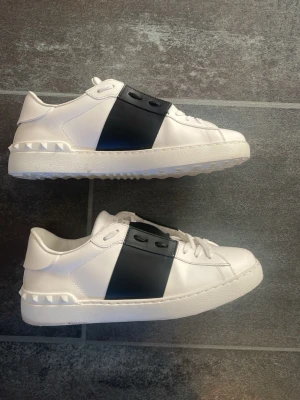 Valentino vita sneakers med svart detalj - Snygga vita sneakers från Valentino med bred svart rem över mitten och coola nitar på hälen. Skorna är i läder och har klassisk låg profil med vita snören. Perfekta för dig som vill ha en stilren men ändå unik look.  Köpta för 4500 hos carlino så 100% autentiska. Helt nya och  rengör även skorna innan de skickas, använda ca 5 gånger. Har ni frågor är de bara ställa!