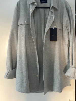 Ljusgrå overshirt från Lindbergh - Stilren ljusgrå overshirt från Lindbergh med klassisk krage, två bröstfickor med knappar och långa ärmar. Aldrig använd, prislapp kvar. 
