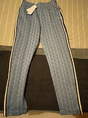 Blå mönstrade mjukisbyxor Lacoste - Säljer ett par blå mjukisbyxor från Lacoste med diskret geometriskt mönster och vita sidoränder. Byxorna har elastisk midja och den klassiska gröna krokodilloggan på benet. Perfekta för en chill och sportig look.