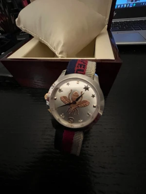 Gucci klocka  - Säljer en Gucci-klocka i originalförpackning. Klockan har ett unikt bi-motiv på urtavlan och ett klassiskt svart och rött armband. Perfekt för både vardag och fest. Kommer med originaldokumentation.