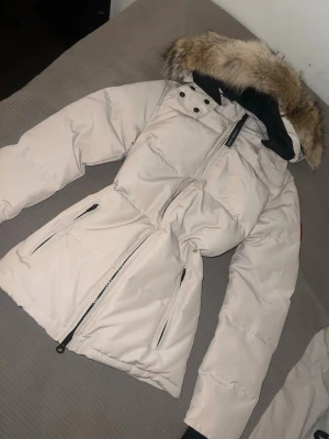 Vit dunjacka från Canada Goose - Snygg vit dunjacka från Canada Goose med stor huva och äkta pälsdetalj. Jackan har två dragkedjeförsedda fickor framtill, svart innerfoder och klassisk Canada Goose-logga på ärmen. Perfekt för kalla vinterdagar och riktigt varm.