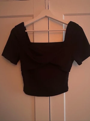 Svart croppad topp med omlott - Säljer min svarta croppad topp med omlottdetalj framtill och korta ärmar. Toppen har en fyrkantig ringning och sitter tight. 