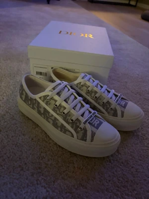 Walkn dior tjej skor - Snygga låga sneakers från Dior med klassiskt grått och vitt Oblique-mönster. Skorna har vita snören, gummisula och detaljer med Dior-logga på tungan och sidan. 