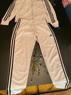Adidas träningsset med svarta ränder - Snyggt träningsset från Adidas med klassiska svarta ränder längs ärmar och ben. Setet består av en zip-jacka med fickor och matchande byxor med dragsko i midjan. Båda delarna har Adidas-logga. Perfekt för sport eller chill.