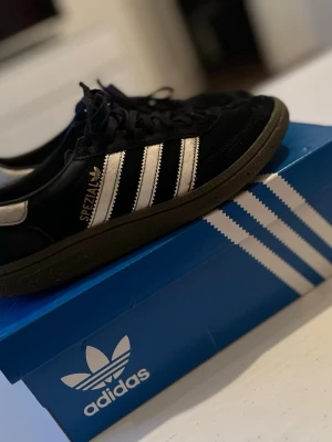 Adidas Spezial svarta sneakers - Säljer ett par Adidas Spezial sneakers i svart mocka med vita detaljer och klassiska tre ränder på sidan. Skorna har låg profil och svart sula, samt guldfärgad Spezial-logga. Perfekta för dig som gillar retrostil och streetwear.
