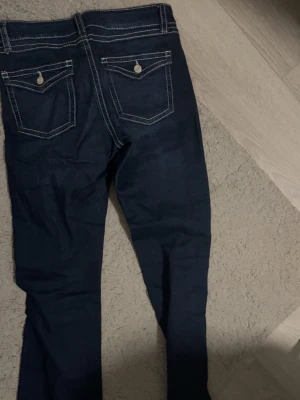Mörkblå jeans med kontrastsömmar - Snygga mörkblå jeans med vita kontrastsömmar och raka ben. Jeansen har två bakfickor med lock och knappdetaljer samt klassisk femficksdesign. Passar perfekt till en avslappnad streetstyle-look.