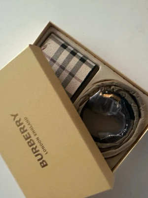 Beige rutigt bälte från Burberry med plånbok - Snyggt beige bälte från Burberry med det klassiska rutiga mönstret i svart, vitt och rött. Bältet har en stilren design och levereras i originalförpackning. Perfekt accessoar för att lyfta din outfit med en lyxig touch.