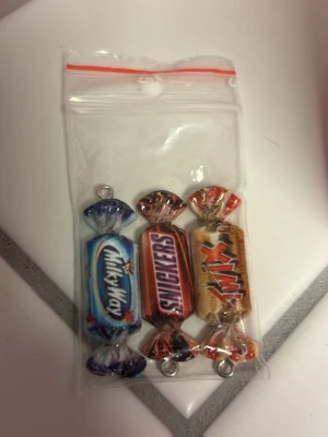 Godisberlocker Milky Way Snickers Twix - Tre färgglada berlocker formade som godisbitar: Milky Way, Snickers och Twix. Tillverkade i plast med metallögla för att fästa på smycken eller nyckelring. Perfekt för dig som gillar söta detaljer och vill pimpa ditt armband, halsband eller väska.