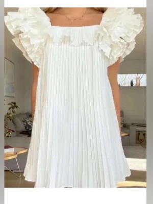 Vit plisserad kortklänning med volang - Superfin vit kortklänning med plisserat tyg och stora volangärmar. Klänningen har en luftig passform och fyrkantig ringning, perfekt för en trendig och somrig look.