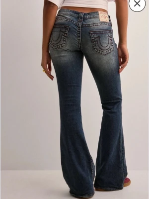 Blå bootcut jeans från True Religion - Säljer ett par blå bootcut jeans från True Religion med ikoniska kontrastsömmar och broderade bakfickor. Jeansen har slitningar och ett hål på ena benet för en trendig, avslappnad look. Klassisk femficksmodell i denim med låg midja.