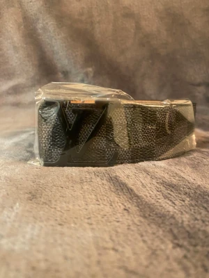 Louis Vuitton svart bälte med LV-spänne - Snyggt svart bälte från Louis Vuitton med klassiskt LV-spänne i svart metall. Bältet har ett diskret mönster och är gjort i skinn med ljus insida. Perfekt accessoar för att lyfta din outfit.