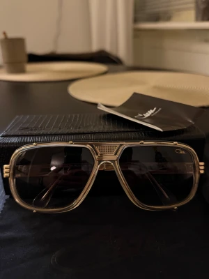 Cazal 665 004 - Cazal modell 665 solglasögon, en designerpärla som kombinerar sofistikering och stil i varje detalj. Den rektangulära ramen i en elegant kristall-guld nyans blandas med utsökt utformade skalmar, vilket ger en touch av distinktion. Den noggrant skulpterade bron säkerställer en bekväm och raffinerad passform. Med en kaliber som anpassar sig till alla ansikten är dessa unisexglasögon det perfekta valet för dem som vill sticka ut med diskret, tidlös elegans.