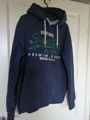 Blå hoodie från Superdry med tryck - Liten missfärgning på snöret som inte märks av. Mörkblå hoodie från Superdry med grönt och vitt tryck på bröstet. Klassisk känguruficka framtill, vita dragsnören i huvan och mjukt fleecefoder på insidan. Perfekt för dig som gillar streetwear och vill ha en skön och snygg hoodie.