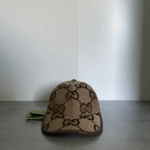 Jumbo Beige Gucci - Säljer en beige keps från Gucci med det klassiska GG-monogrammet i brunt över hela kepsen. Kepsen har böjd skärm och bruna detaljer längs kanterna. Perfekt för dig som vill ha en stilren accessoar med lyxig känsla. Aldrig använd. Storlek M 58cm.