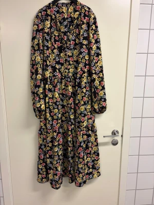 Blommig långklänning med volangfrpn rut&circle - Långklänning med svart botten och färgglatt blommönster i gult, rosa och lila. Klänningen har långa ärmar med resår vid ärmslut och volangdetaljer längs kjolen. Luftig passform och v-ringad hals. Perfekt för dig som gillar romantisk stil.
