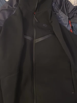 Svart Nike hoodie med dragkedja - Snygg svart hoodie från Nike med hel dragkedja framtill och diskreta loggor på bröstet och ärmen. Hoodien har huva och detaljer i mörkgrått samt sportig design. Perfekt för dig som gillar streetwear och sportig stil.