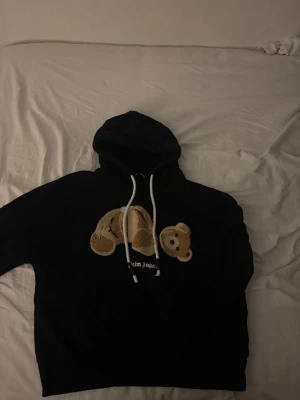 Svart hoodie Palm Angels med björn - Svart hoodie från Palm Angels med huva och vita dragsnören. På bröstet finns ett broderat motiv av en nallebjörn med avtagbart huvud i beige och brunt. Hoodien har en avslappnad passform och är tillverkad i mjukt bomullsmaterial.