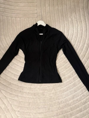 Svart ziptröja med hög krage - Snygg svart ziptröja med figurnära passform och hög krage. Tröjan har långa ärmar och dragkedja framtill, perfekt för en sportig och stilren look. Passar till många olika outfits och är enkel att matcha.