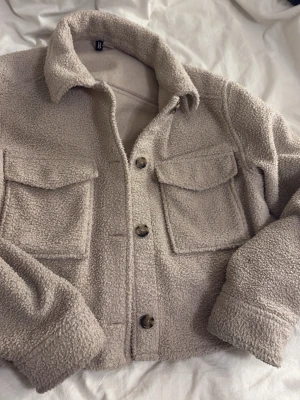 Beige teddyjacka med stora fickor - Supermjuk och fluffig teddyjacka i beige med två stora bröstfickor och knappar framtill. Jackan har en klassisk krage och så har ena knappen gått av men de går att fixa, perfekt inför hösten Materialet är teddy/fleece som ger en mysig känsla. Passar både M och S